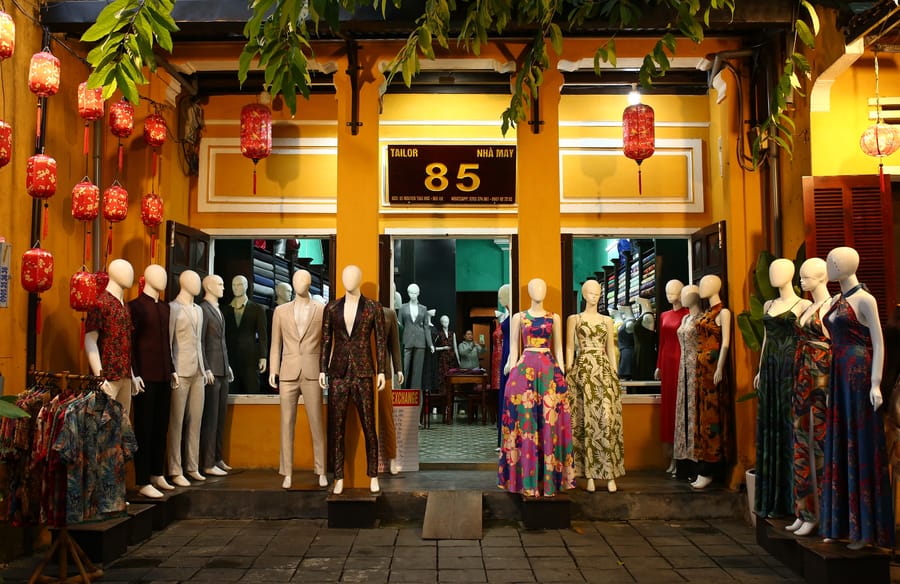 A Hội An tailor shopfront at night, its entrance lined with mannequins in suits and dresses beneath rows of red lanterns.