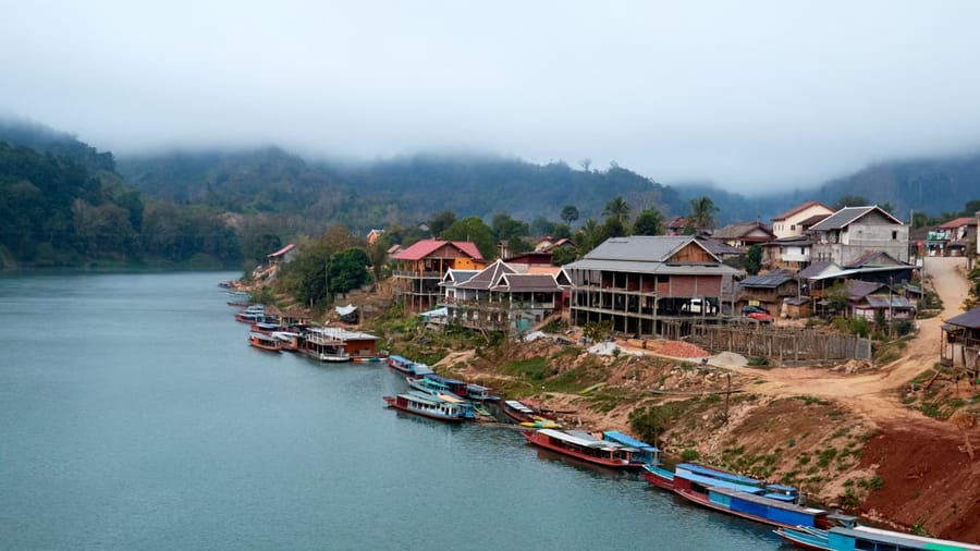 A riverside Lao village of wooden stilt houses with long-tail boats moored along the bank, forested hills fading into mist behind.