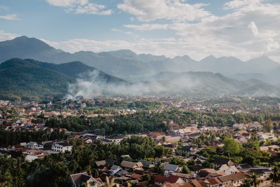 An elevated view over Luang Prabang’s rooftops and palm trees, with hazy mountains and rising smoke behind the town.