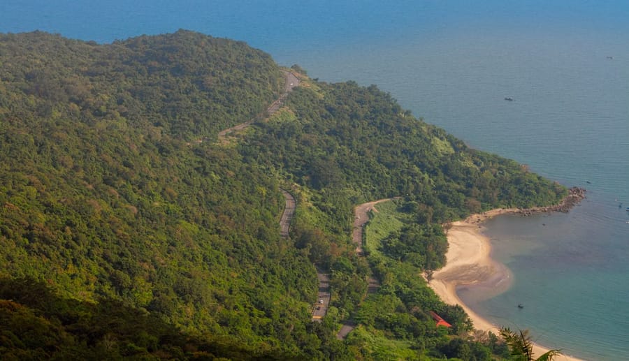 An aerial view of a winding mountain road cut through jungle along the coast, dropping toward a small sandy cove.