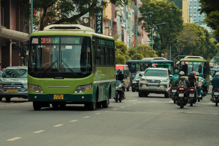 A green public bus and a stream of cars and motorbikes moving down a busy city street lined with shops.