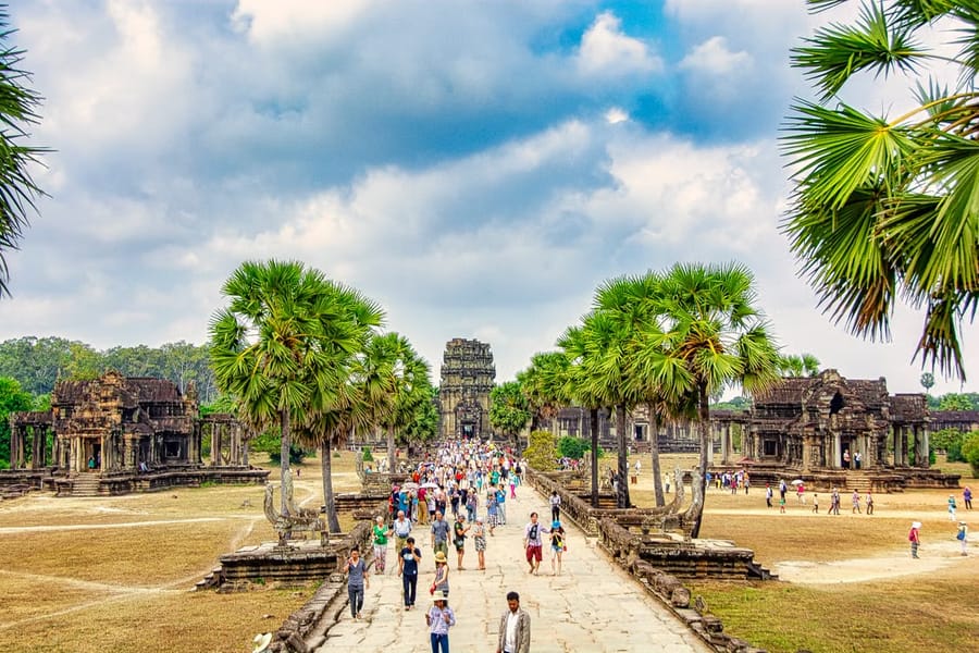 Crowds of tourists walking the long stone causeway to Angkor Wat, lined with sugar palms leading to the temple’s towers.