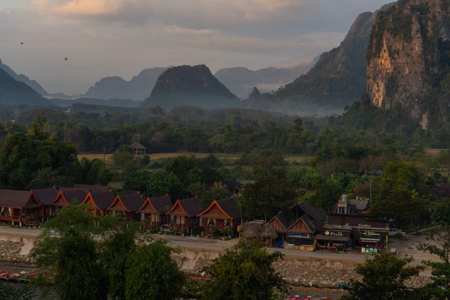 Riverside bungalows in Vang Vieng at dusk, with hot-air balloons drifting over the surrounding limestone karst mountains.