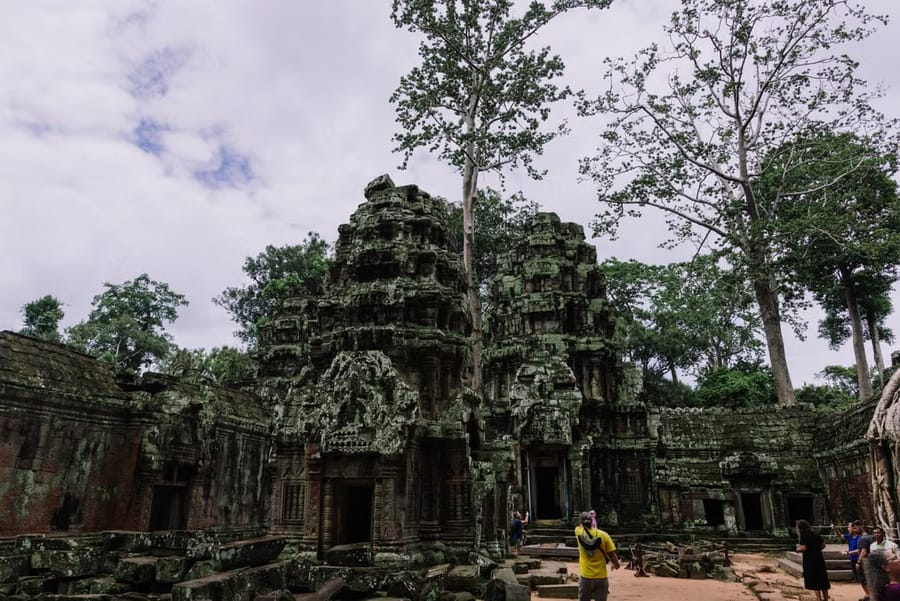 Moss-covered Angkor-style temple towers overgrown with jungle trees, with a few visitors exploring the ruins below.