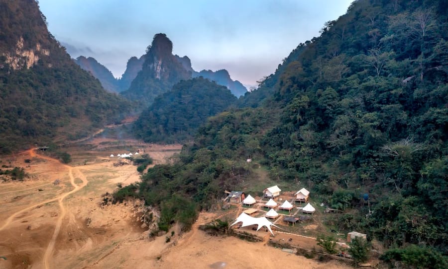 A cluster of white tents sits on a dry riverbed in a forested Lao valley, framed by jagged karst peaks.