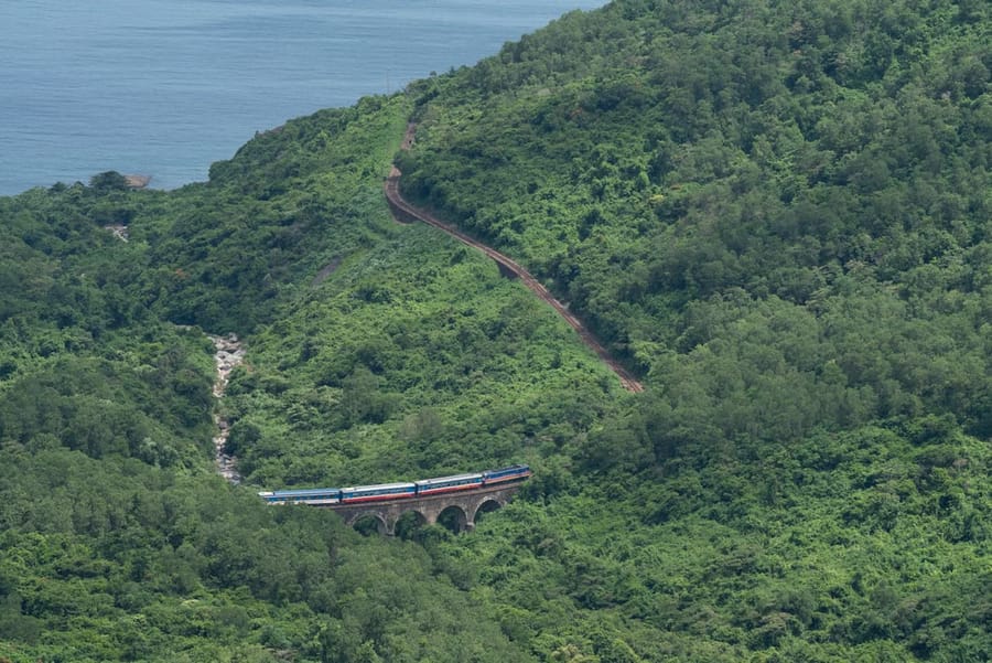 A Reunification Express train crosses a small stone viaduct on a forested mountainside above the coast.