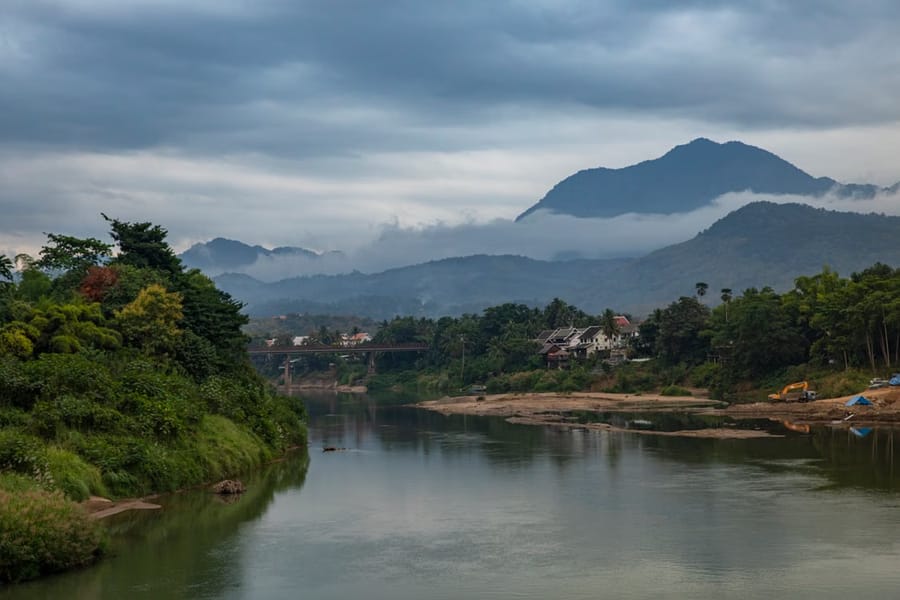 A calm river winds through a misty, mountain-ringed valley in Laos, with a small bridge crossing it in the distance.