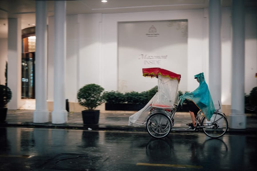 A cyclo driver in a rain poncho pedals an empty, plastic-covered cyclo past a hotel colonnade on a wet street at night.