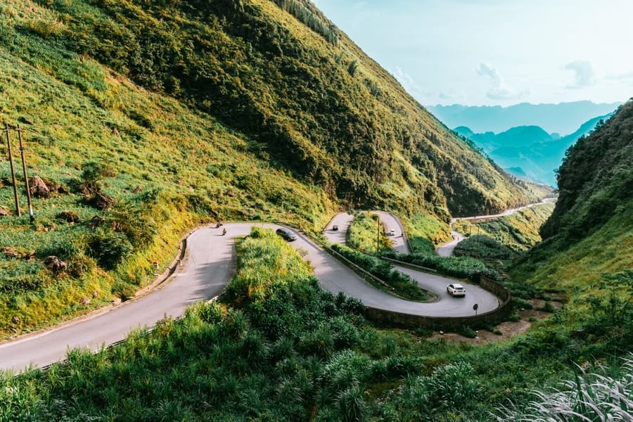 A road coils in hairpin bends up a green mountainside on the Hà Giang Loop, with a car and motorbikes climbing towards distant blue ridges.
