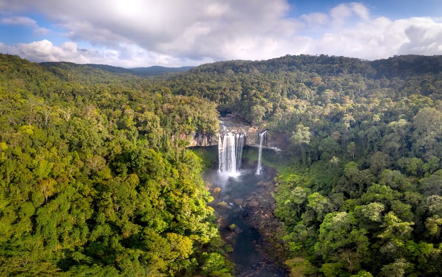 Seen from the air, a waterfall drops in two white streams into a rocky gorge surrounded by dense green forest.