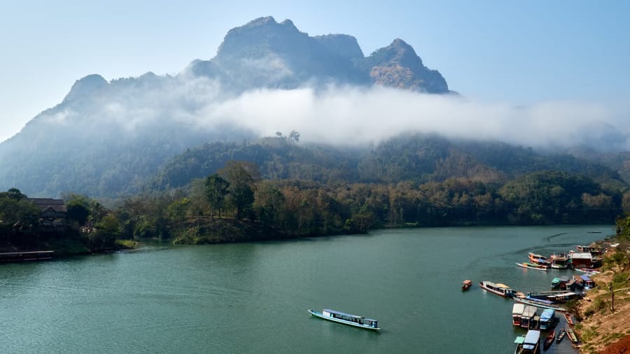Mist clings to a jagged limestone peak above the Nam Ou river at Nong Khiaw, where long-tail boats wait along the bank.